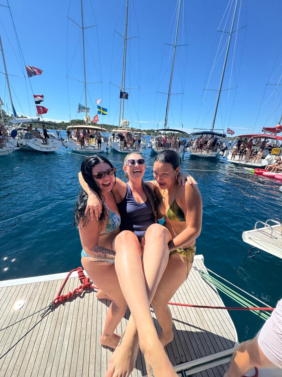 Three women sitting on the bow of a yacht with their feet over the water, other yachts rafted alongside