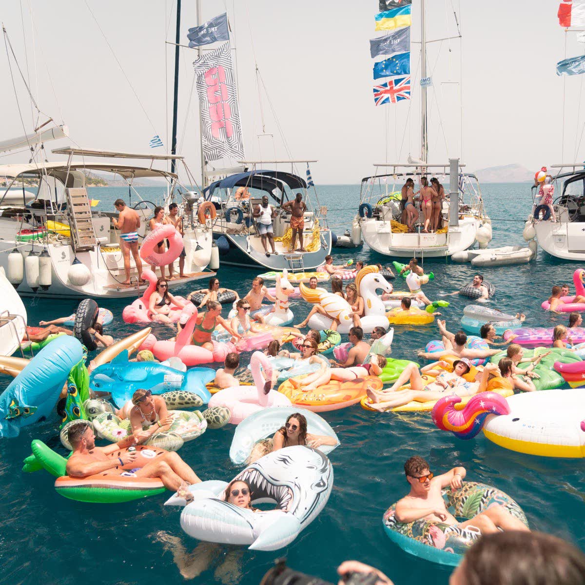 A crowd on inflatables in the water between rafted yachts, flags flying overhead