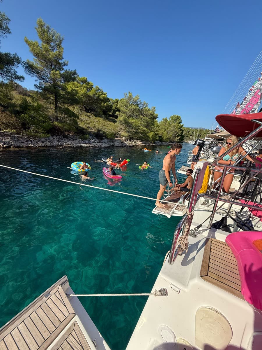 Looking down the side deck of a yacht into a green bay, people floating on inflatables in clear water