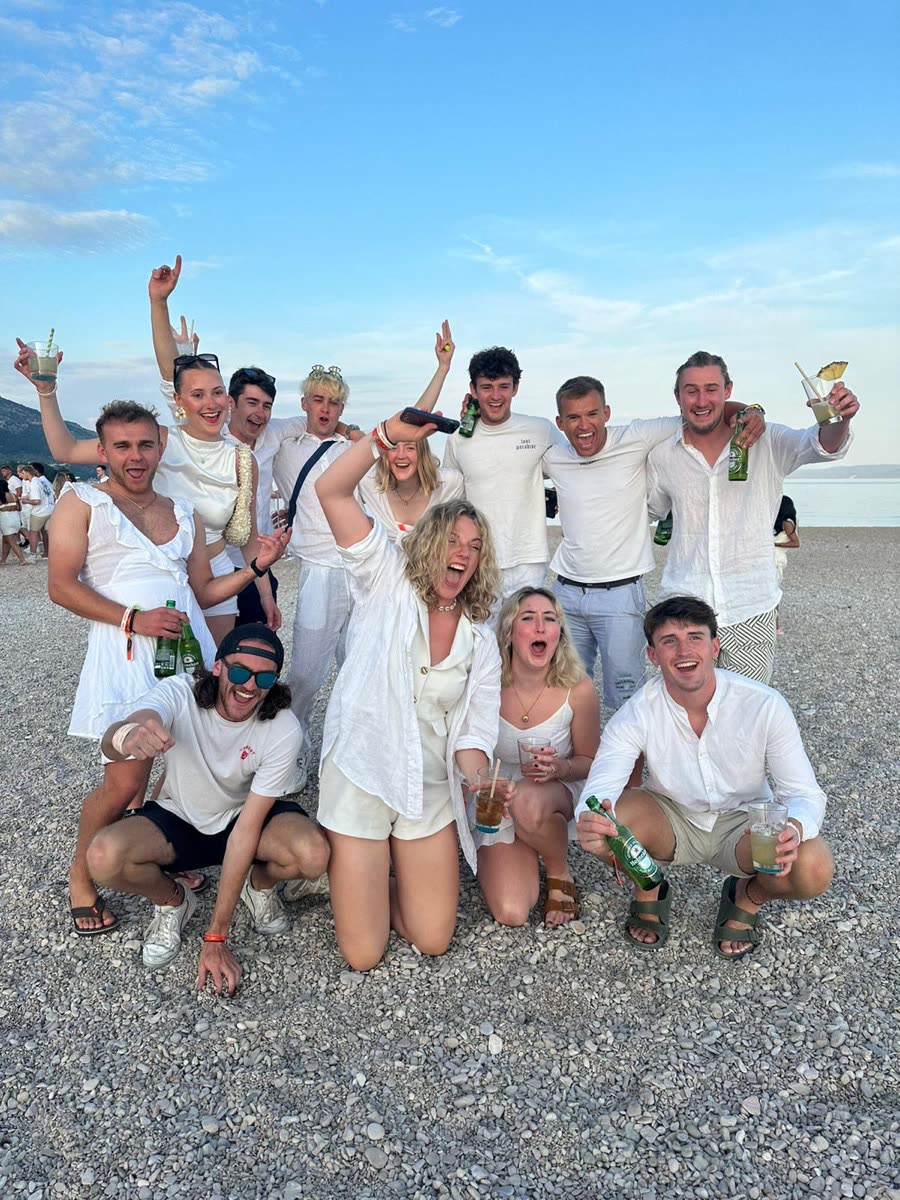 A group in white on a pebble beach at dusk, cheering with drinks in the air