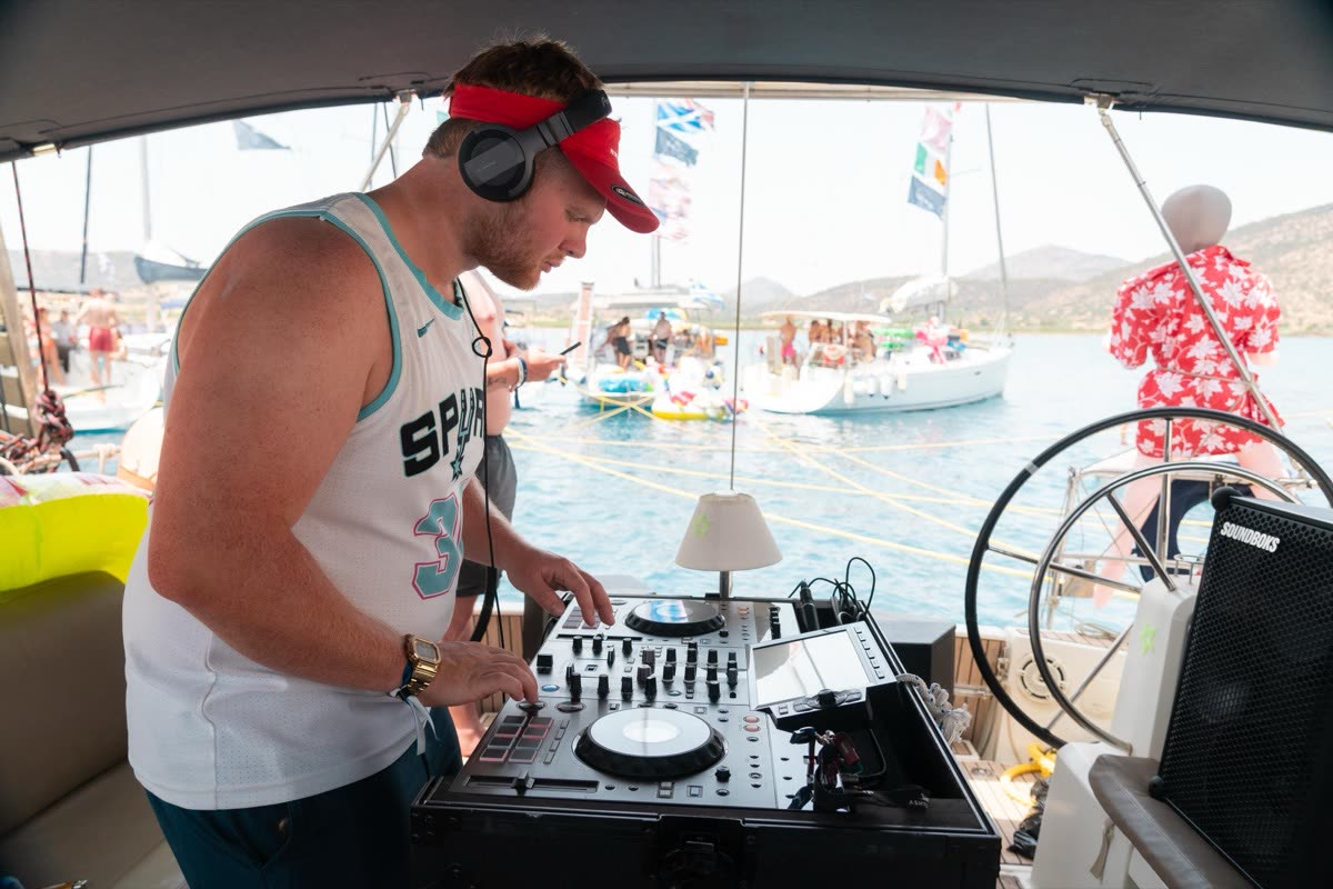 A DJ in a cap and headphones playing a set on the back of a yacht, the rafted fleet behind