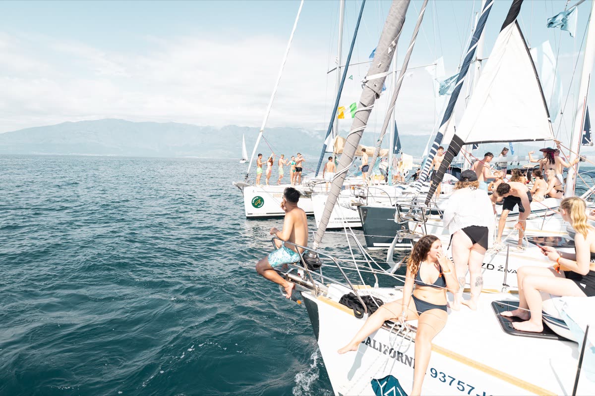A yacht under sail with people sitting along the deck, more of the fleet behind