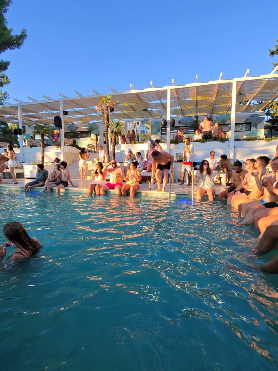 A packed beach club pool with white canopies and people sitting along the edge
