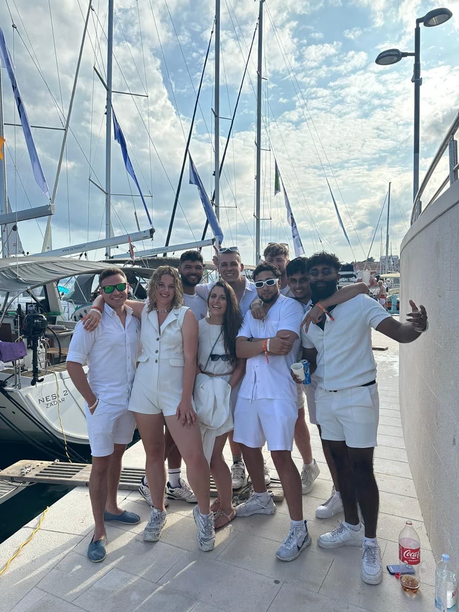 A group dressed in white posing together on a pontoon with masts behind