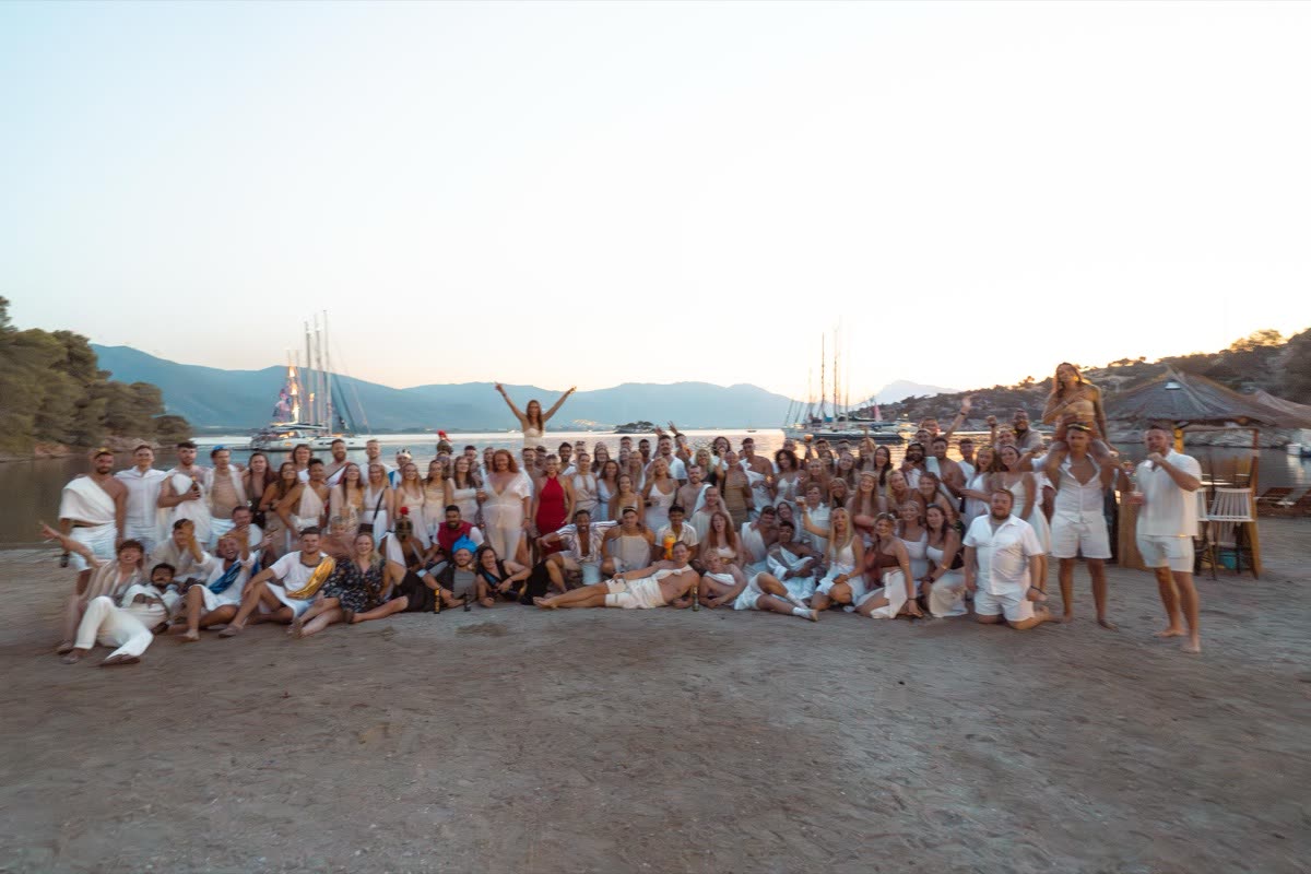 The full group lined up on a beach at dusk with the yachts moored behind