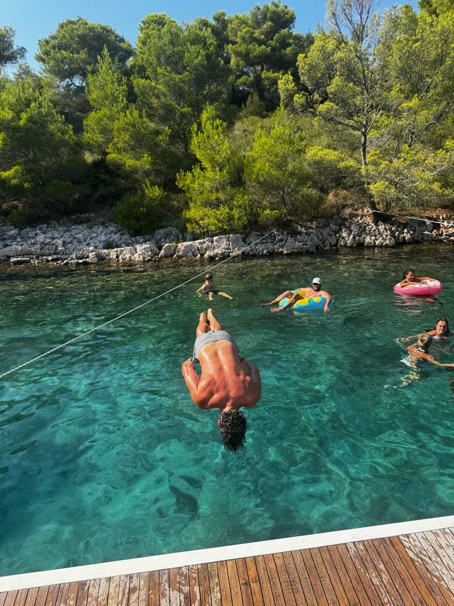 Someone mid dive off a wooden jetty into clear turquoise water, pine trees on the shore behind