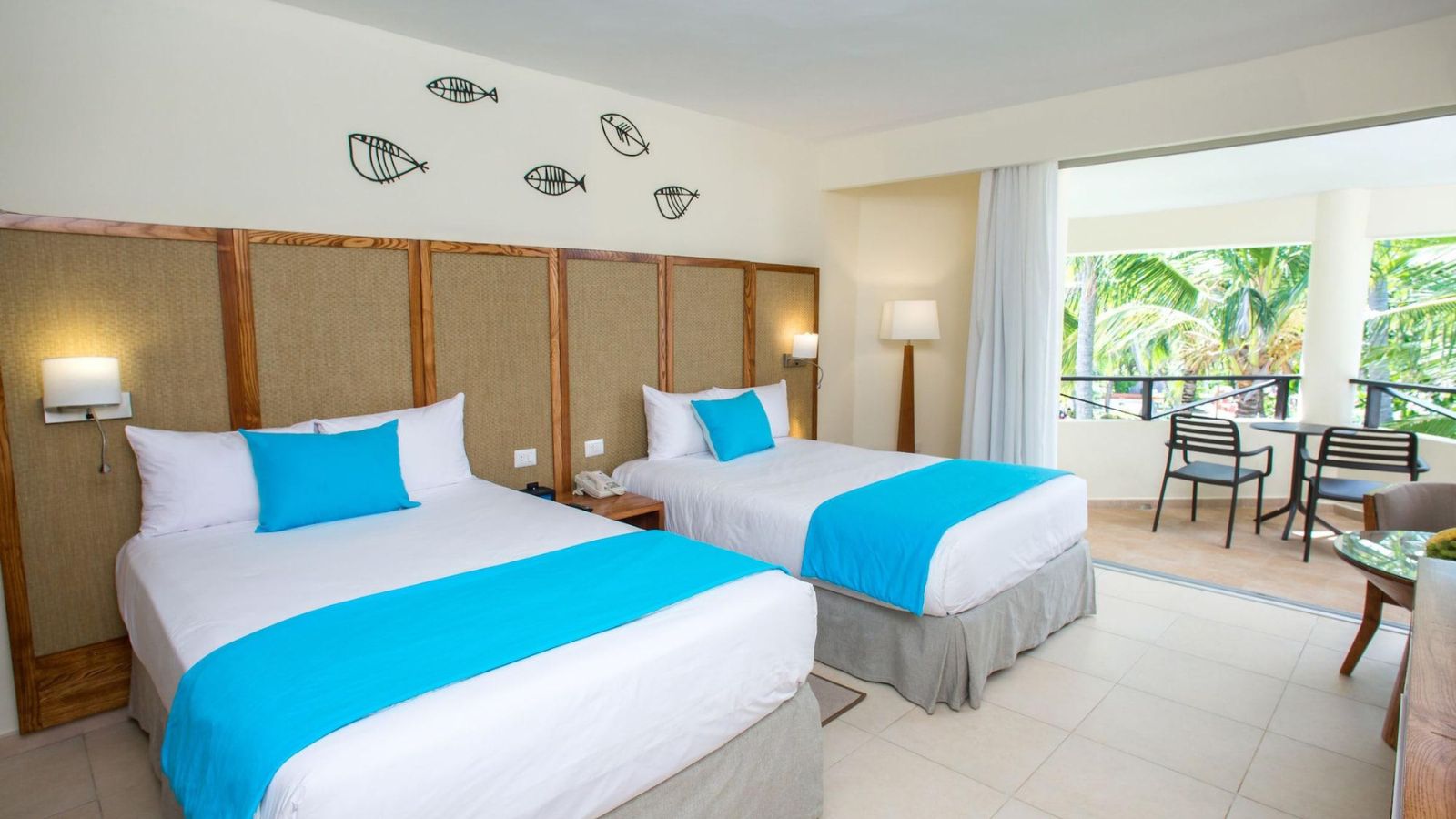 A room with two double beds in white linen with bright blue runners, woven headboard panels and fish wall art, with doors open onto a balcony surrounded by palms