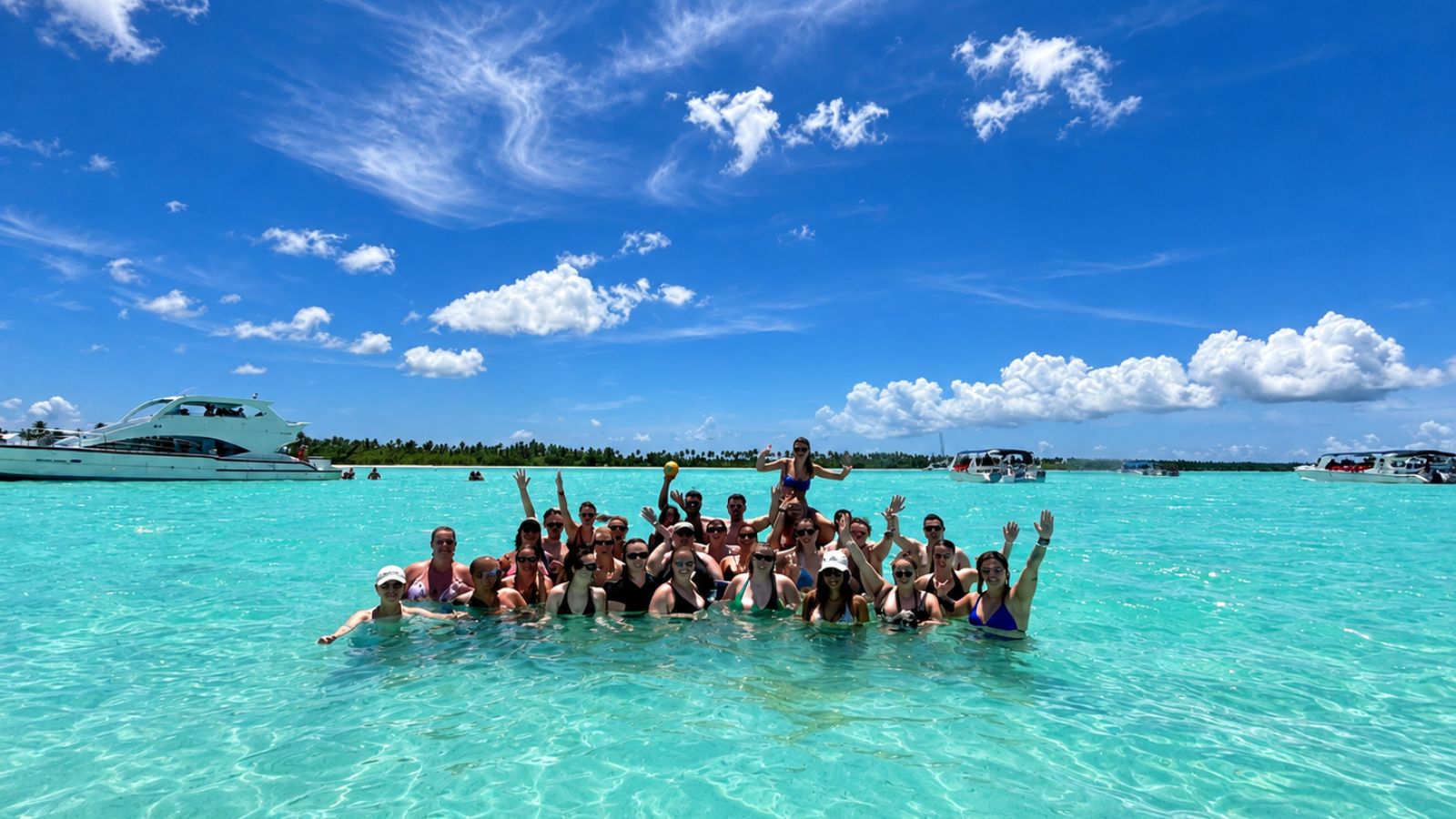 The group standing waist deep in turquoise shallows with arms raised, boats moored behind them and a palm lined shore on the horizon