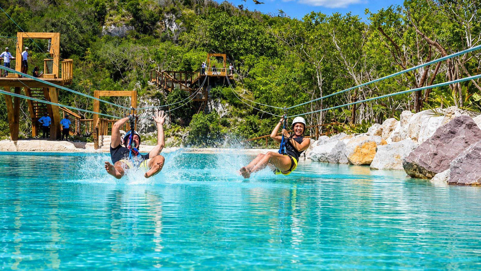 Two people splashing feet first into a turquoise pool at the end of a zip line, with wooden platforms behind