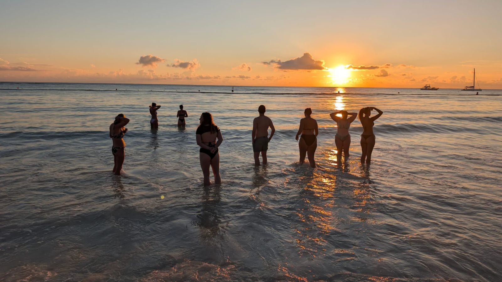 The group silhouetted standing in the shallows as the sun sets over the sea, with a catamaran on the horizon