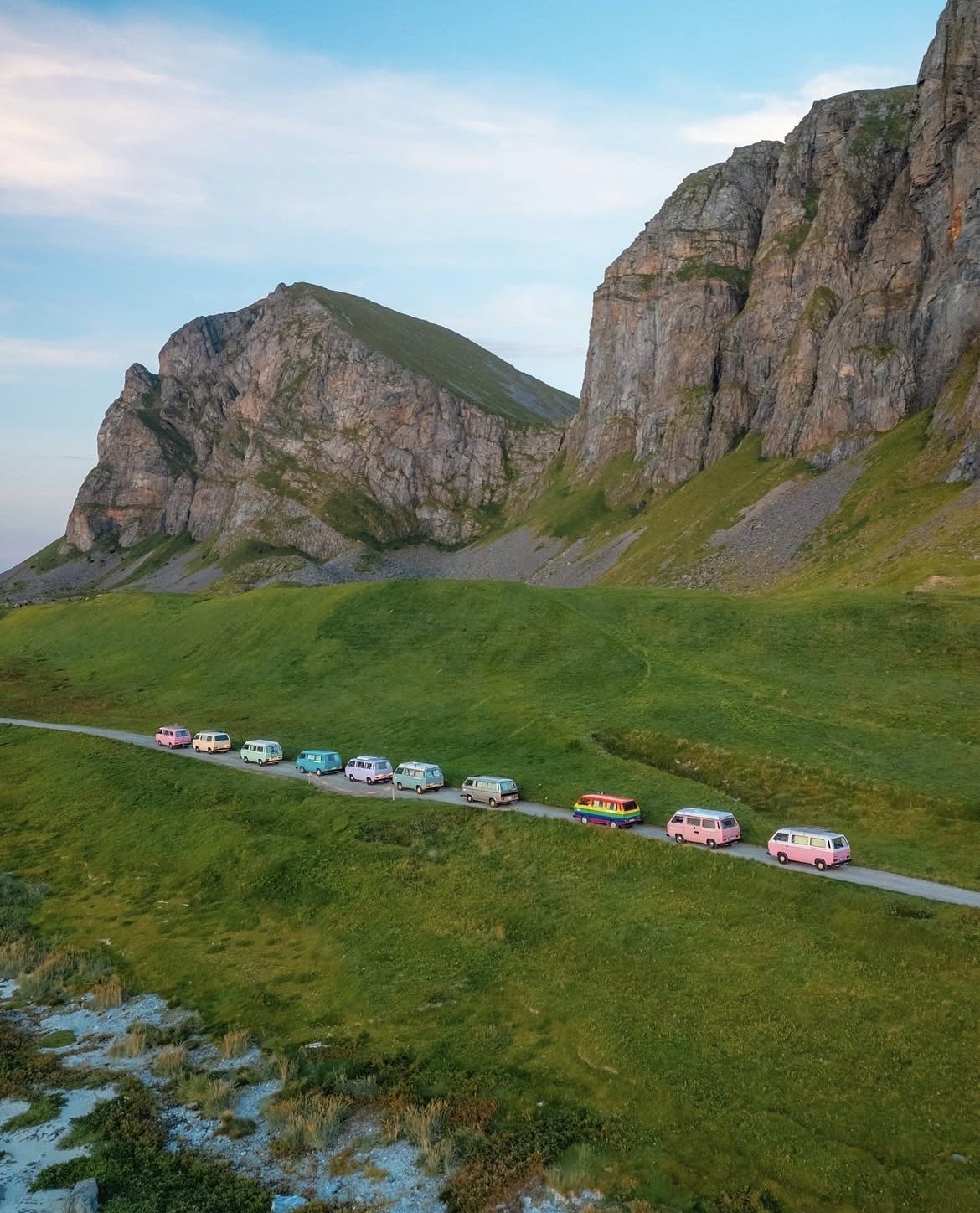 Eleven pastel VW campers nose to tail on a single track road beneath a rock face