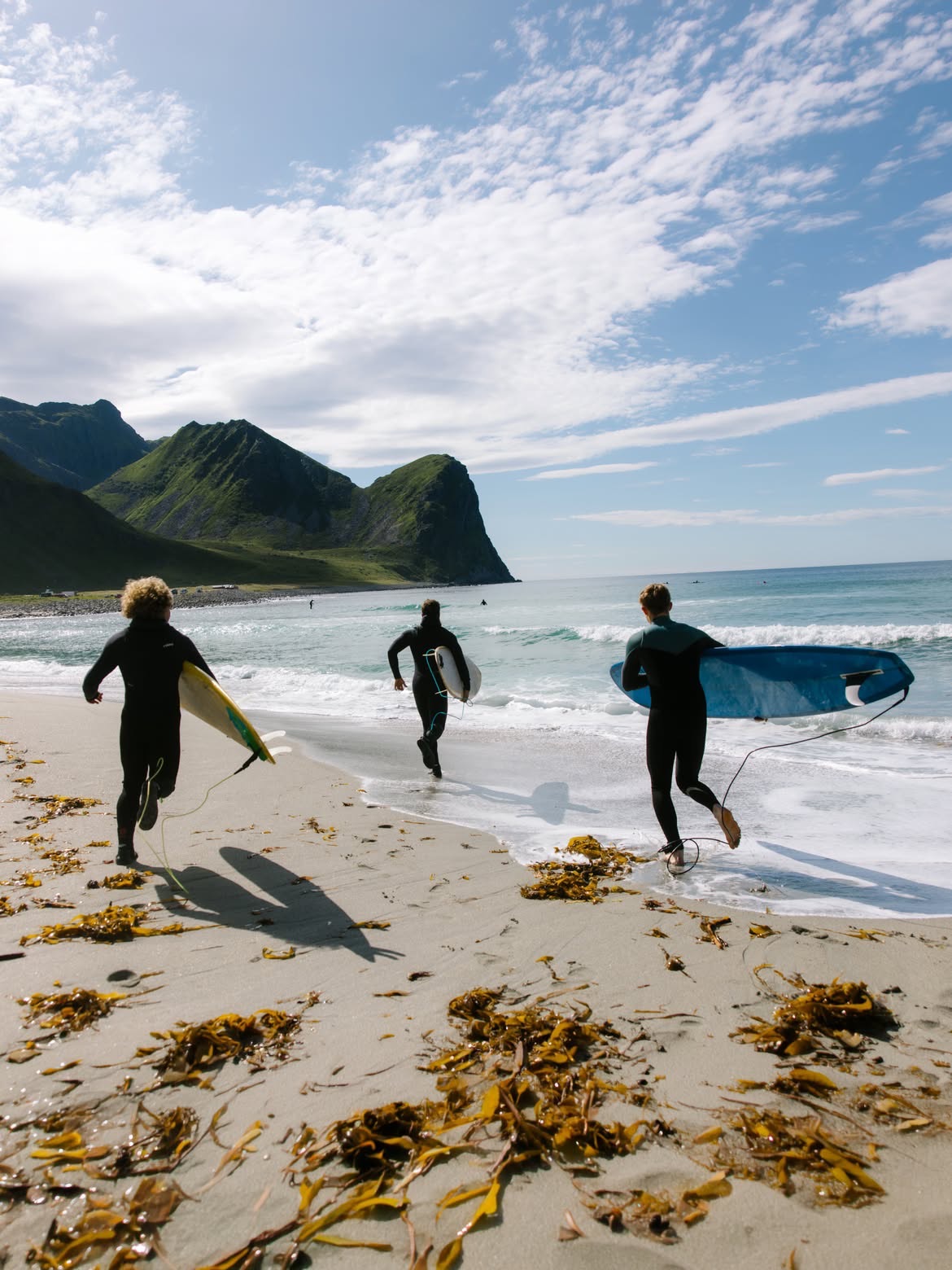 Three surfers running into the water with their boards, kelp across the sand