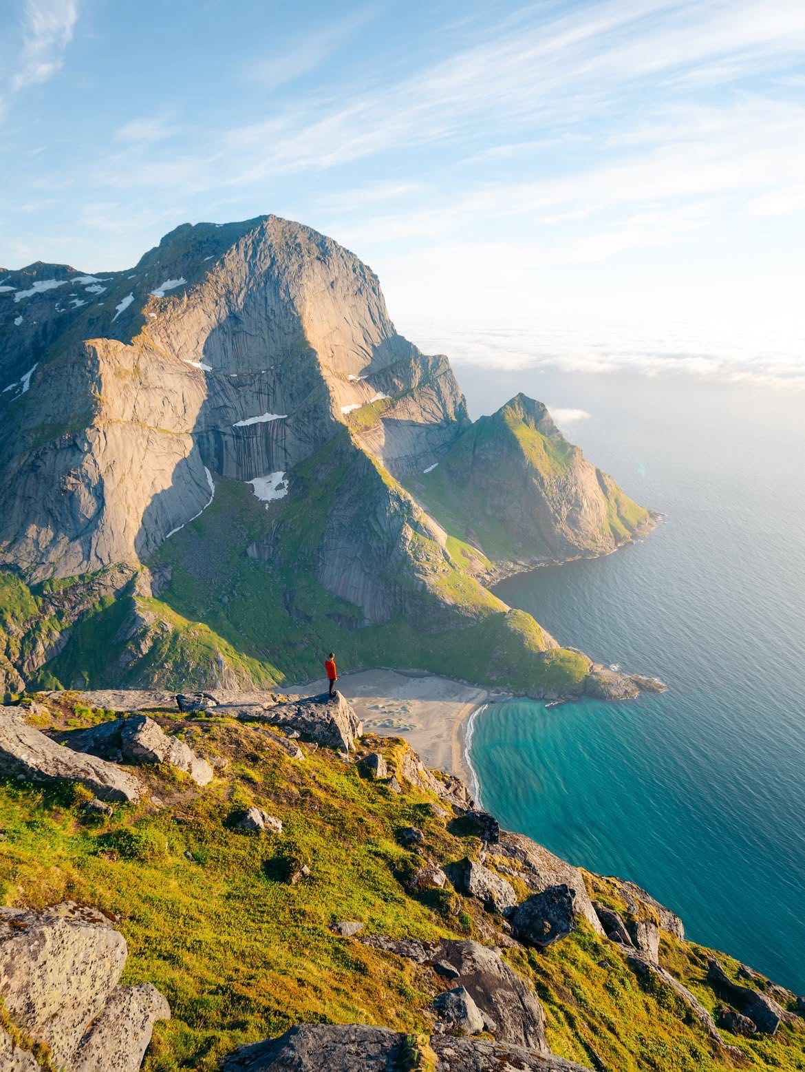 A hiker in a red jacket on a clifftop looking down to a beach far below