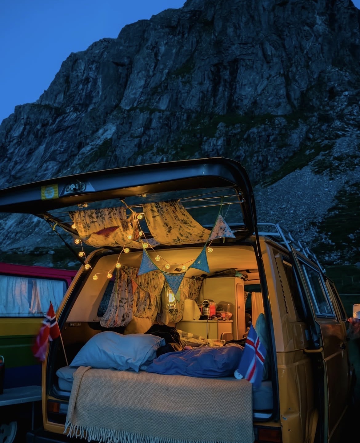 The inside of a camper at dusk, bed made up, fairy lights and bunting