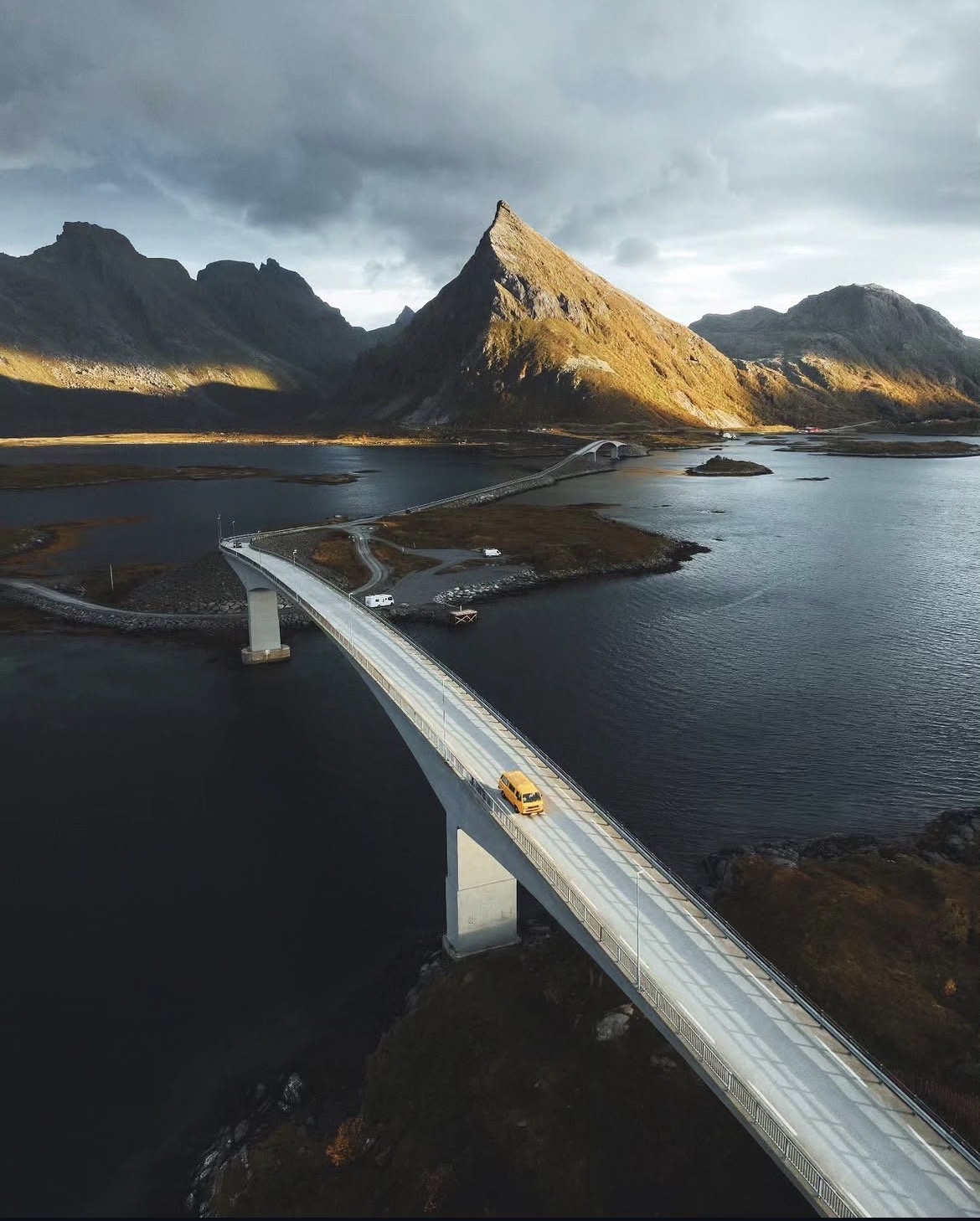 A yellow camper crossing the Fredvang bridge with a pointed peak behind
