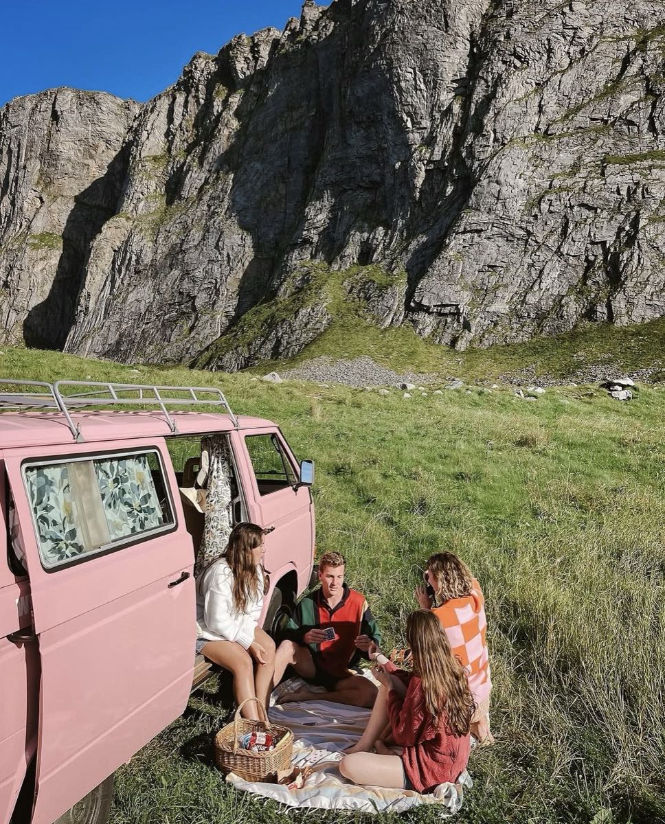 Four people playing cards on a blanket beside a pink camper