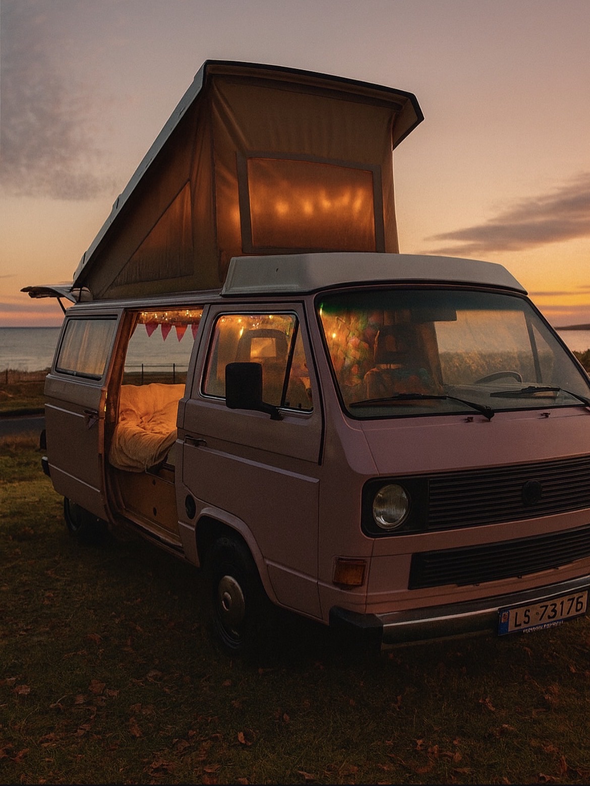 A pink camper with its pop top raised, bed and fairy lights inside, at sunset