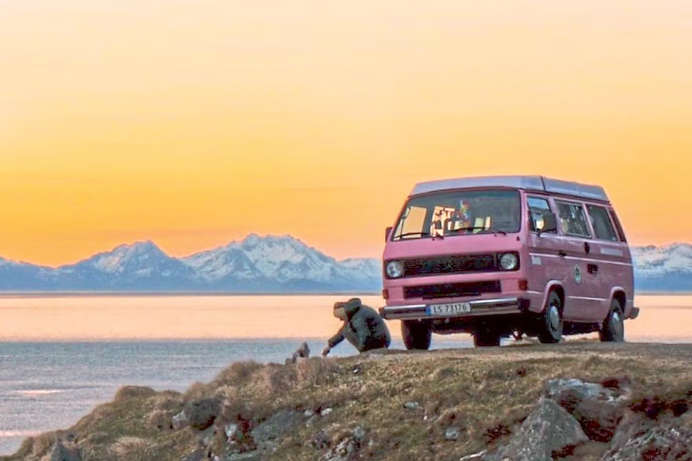 A pink camper parked on a headland with snow covered peaks across the water