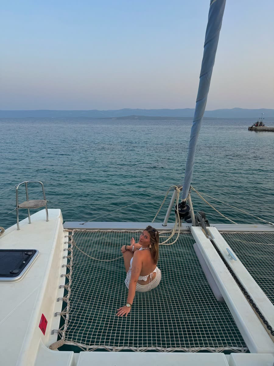 Sitting on the trampoline net between the twin hulls of a catamaran at dusk, flat water and hills on the horizon