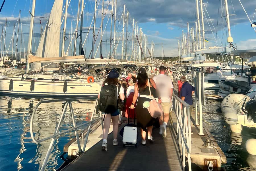 The group walking along the pontoon with their luggage, masts on both sides under a blue sky