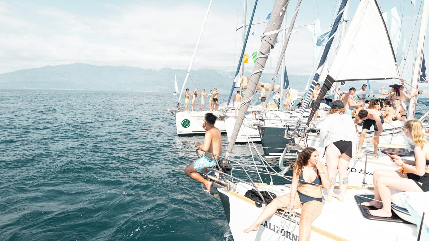 Yachts rafted together on open water, people sitting along the decks and one person climbing down the side into the sea