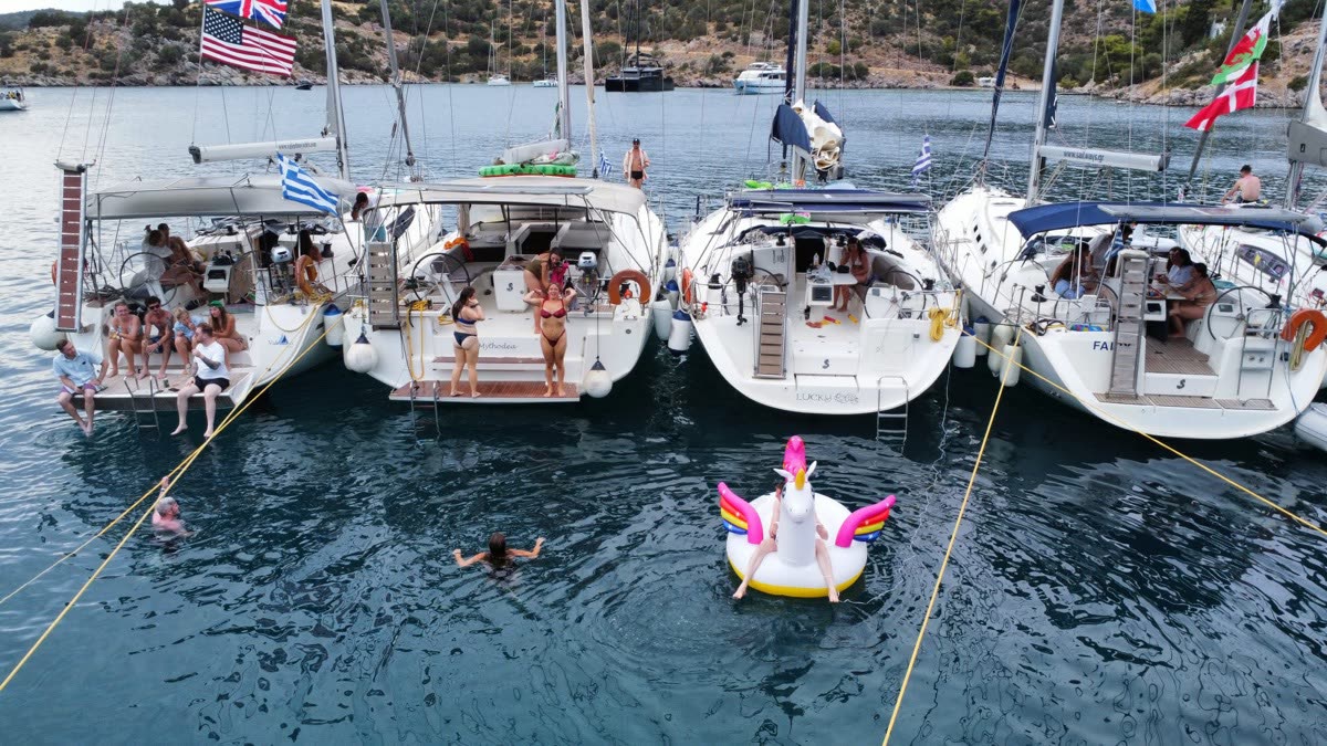 Four sailing yachts rafted stern to stern in a bay seen from above, people along the decks and a unicorn float in the water