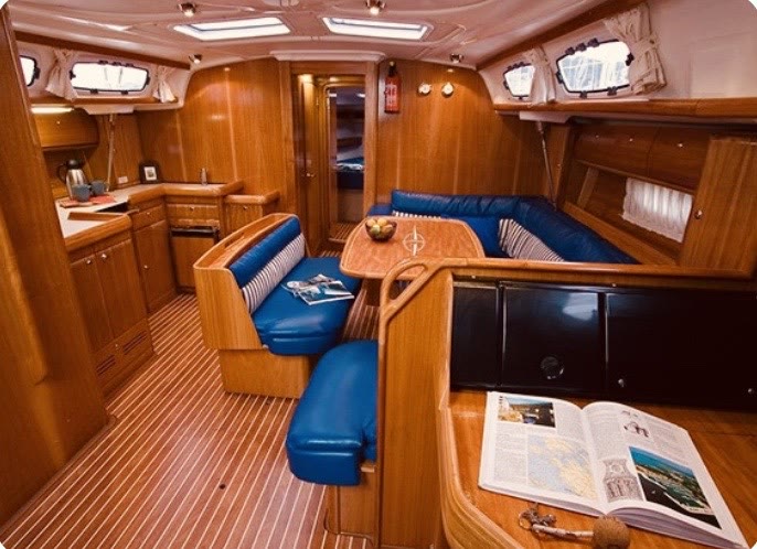 The saloon of a sailing yacht, teak panelling with blue bench seating around a table and a galley along one side