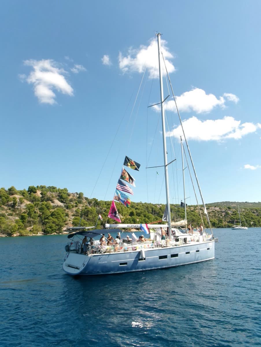 A sailing yacht under way with bunting strung up the forestay, a pine covered shore behind